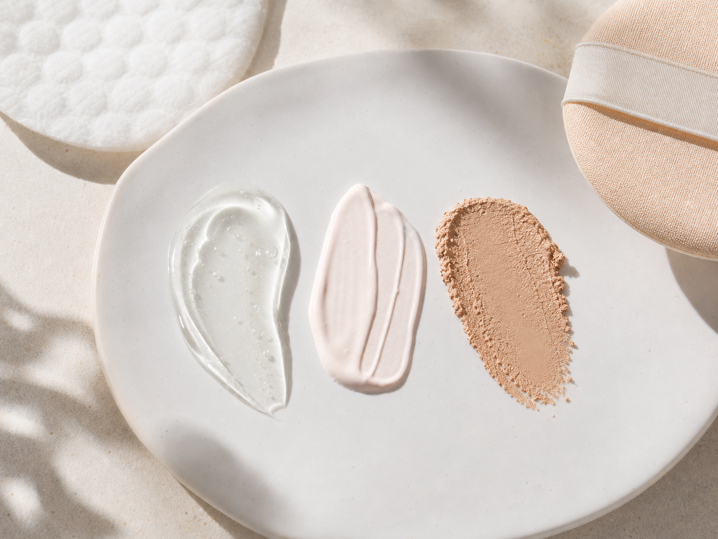 선크림 젤, 톤업 베이스, 쿠션 질감을 흰 접시 위에 나란히 놓은 texture flatlay