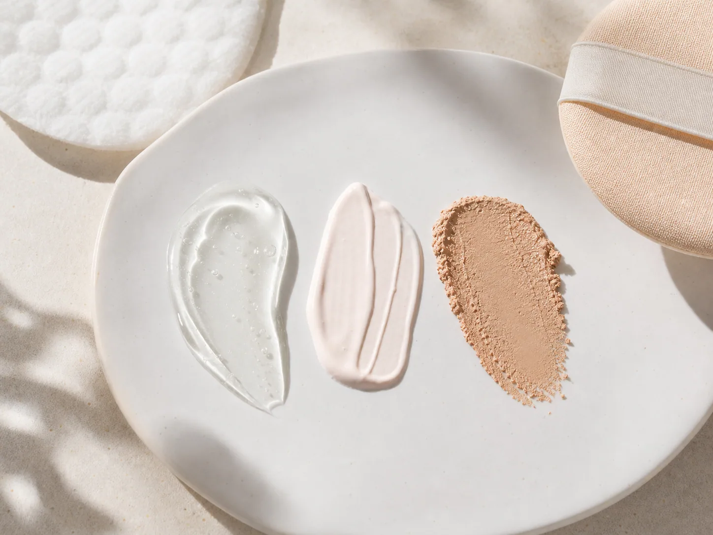 선크림 젤, 톤업 베이스, 쿠션 질감을 흰 접시 위에 나란히 놓은 texture flatlay